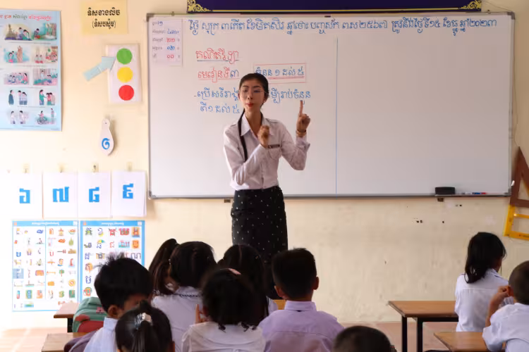 Teacher in a classroom writes on a whiteboard while students sit attentively.