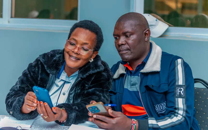 Two people sit closely together, smiling as they look at smartphones. 