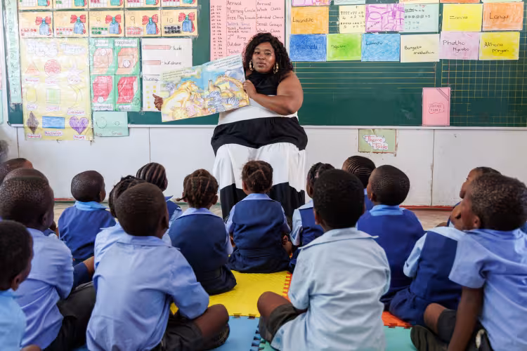 A teacher sits in front of a blackboard, showing a picture book to a group of attentive young students in blue uniforms. Classroom posters decorate the background.