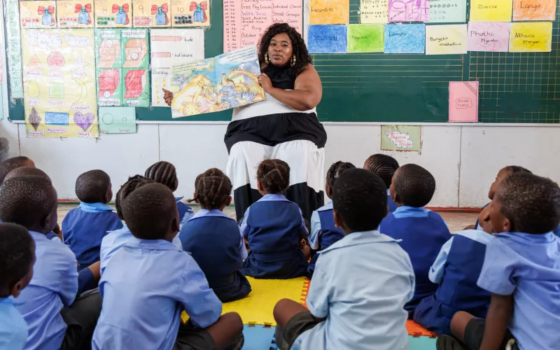 A teacher sits in front of a blackboard, showing a picture book to a group of attentive young students in blue uniforms. Classroom posters decorate the background.