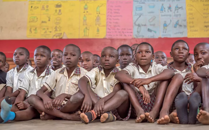 A group of attentive children sit on a classroom floor, wearing checkered uniforms. Colorful educational posters cover the walls, creating a lively atmosphere.