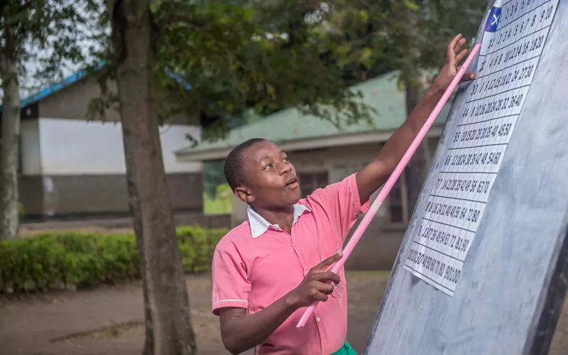 A student wearing a pink shirt and green shorts points eagerly with a stick at a chart on an outdoor blackboard, surrounded by trees and a building.