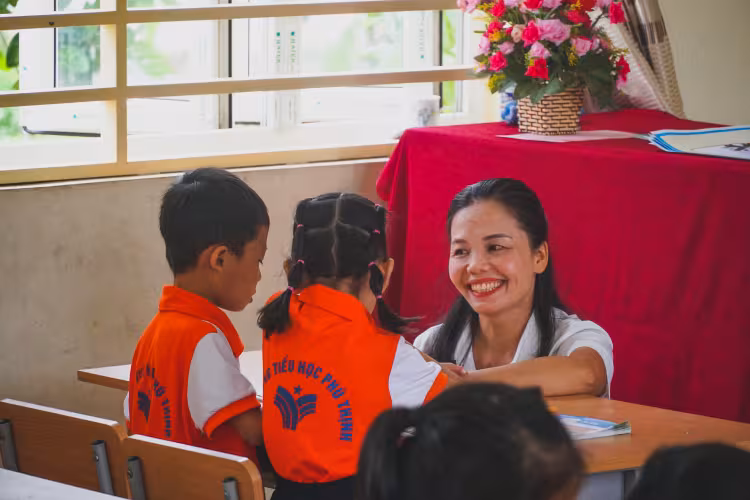 A teacher smiles while interacting with two children in orange uniforms at a school desk.