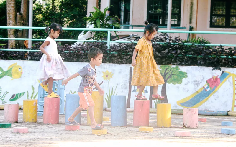 Three children playfully balance on colorful stepping stones in a sunny playground, backed by a mural of nature and a cheerful scene.