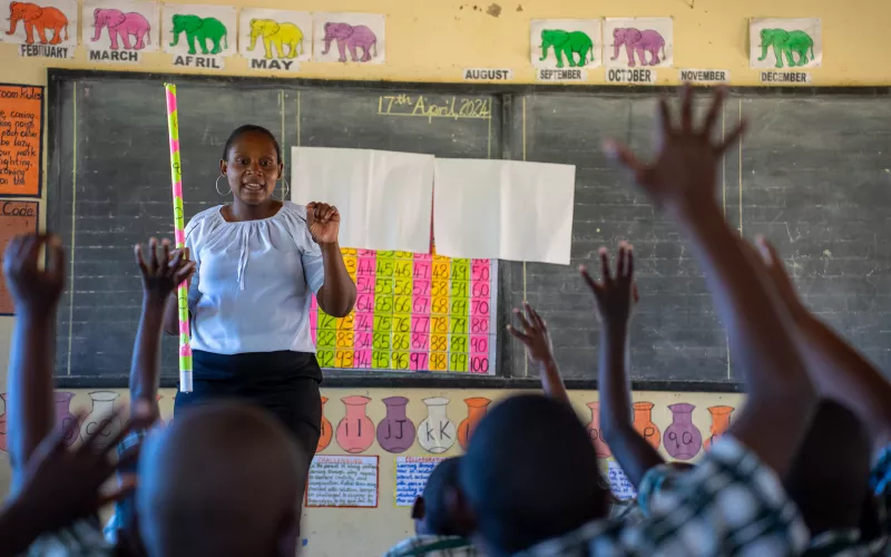 A teacher in a Zambian classroom stands in front of a chalkboard, while students raise their hands to answer.