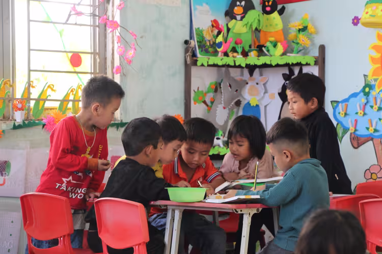 A group of young children sit around a table in a colorful classroom, engaged in an activity.