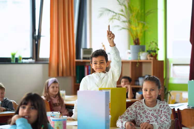 A young boy in a classroom eagerly raises his hand, surrounded by attentive classmates. The room is bright, with plants and colorful dividers.