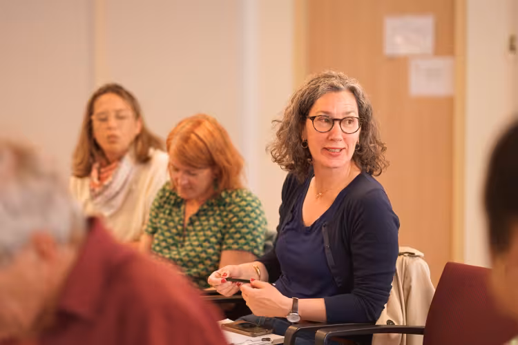 A woman with curly hair and glasses attentively listens in a meeting, surrounded by others. The room conveys a focused and engaged atmosphere.