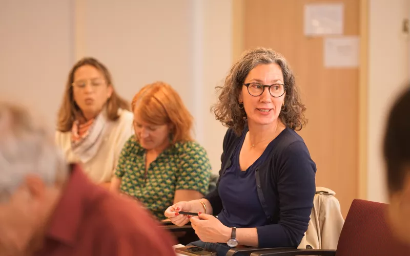A woman with curly hair and glasses attentively listens in a meeting, surrounded by others. The room conveys a focused and engaged atmosphere.