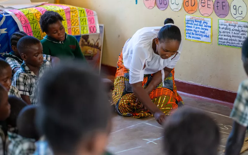 A teacher in a Zambian classroom writes on the floor with chalk, surrounded by learners.