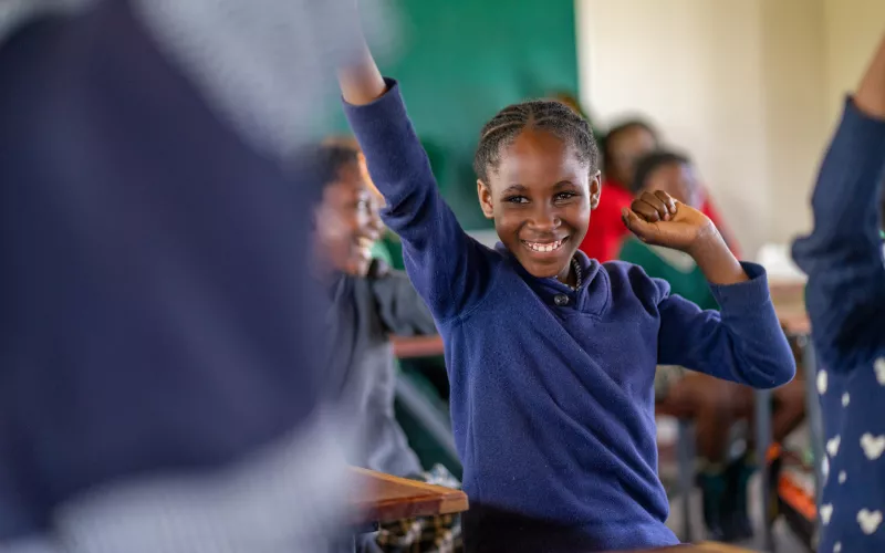 A Zambian schoolgirl in a navy uniform raises her hand, smiling.