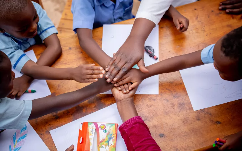 Six children's hands come together over a desk to demonstrate teamwork