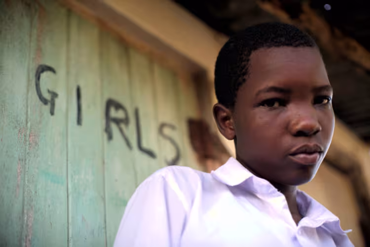 A young black girl in a white shirt stands in front of a weathered green door with "GIRLS" painted on it, looking contemplative and serious.