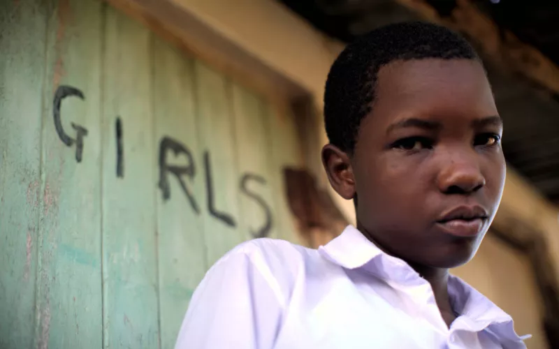 A young black girl in a white shirt stands in front of a weathered green door with "GIRLS" painted on it, looking contemplative and serious.