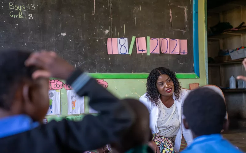Teacher Emelda sits on the floor at the front of a classroom, speaking to a group of young children facing her.