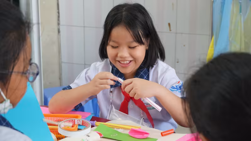Smiling girl in a school uniform engages in arts and crafts at a table, surrounded by colorful paper and supplies, with two classmates nearby.