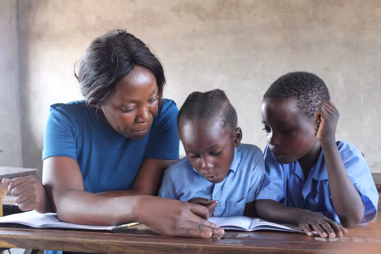 A teacher sits with two children in uniforms at a desk, pointing at a book. The children focus intently, creating a scene of learning and guidance.