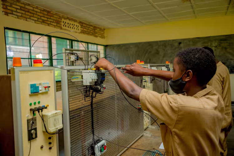 A student wearing a uniform and mask works on electrical wiring in a classroom with equipment panels, conveying focus and technical learning.
