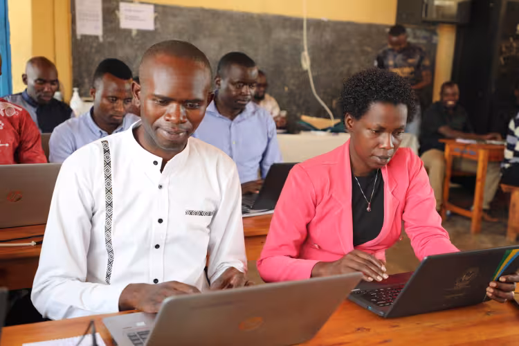 A group of people in a classroom setting, focused on laptops. A man in a white shirt and a woman in a pink jacket are in the foreground. The mood is attentive and studious.