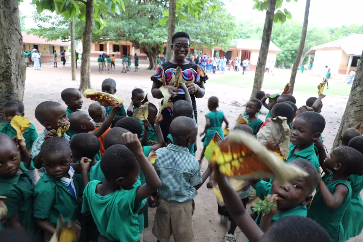 A joyful teacher is surrounded by smiling schoolchildren holding leaves in a lively outdoor schoolyard. The scene conveys happiness and learning.