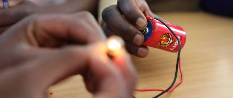 Hands hold a lit bulb connected by wires to a red battery, demonstrating a basic electric circuit. The atmosphere is educational and hands-on.