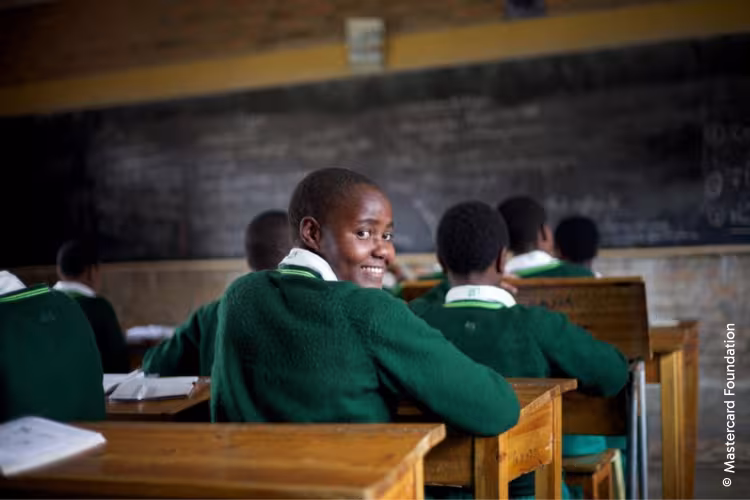 A student in a classroom wearing a green uniform turns and smiles at the camera, surrounded by classmates. A blackboard is in the background.