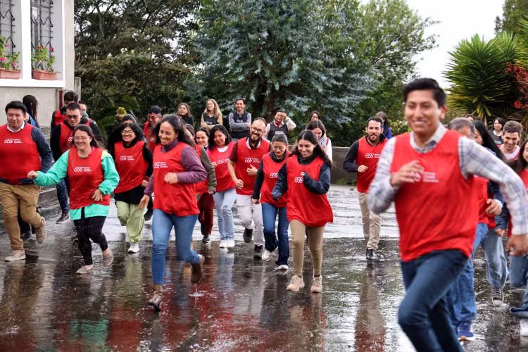 A group of people wearing red vests joyfully run outdoors on a rainy day. They are smiling and surrounded by greenery, creating an energetic atmosphere.