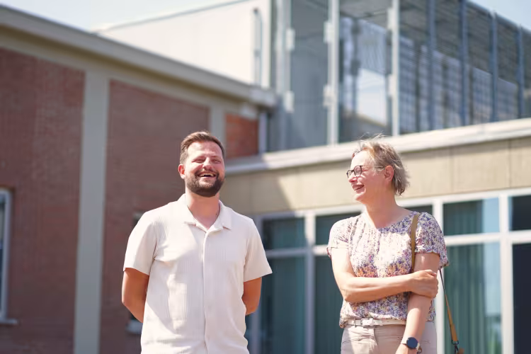 A man and woman are laughing outdoors on a sunny day, standing in front of a modern building with brick and glass elements. The mood is joyful.