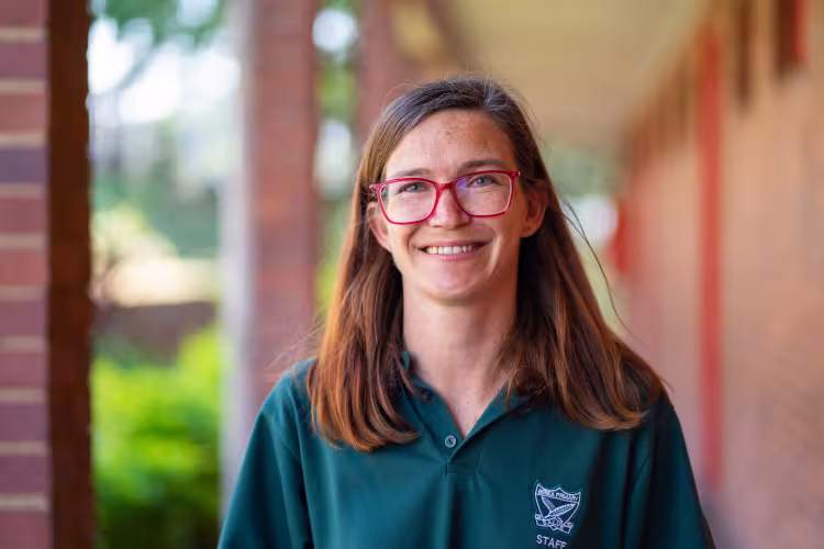 A smiling person wearing a green staff polo shirt stands outdoors along a covered walkway.