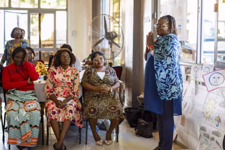 Women in colorful African clothing sit attentively as a woman stands with palms together in a bright community room displaying drawings.
