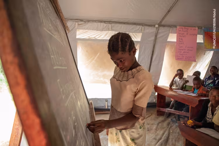 A schoolgirl writes on a chalkboard in a tent classroom as classmates watch from wooden desks, creating a focused, hopeful learning atmosphere.