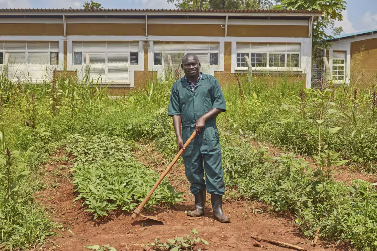 A man wearing green work overalls and rubber boots stands holding a hoe in a vegetable garden in front of a school building.