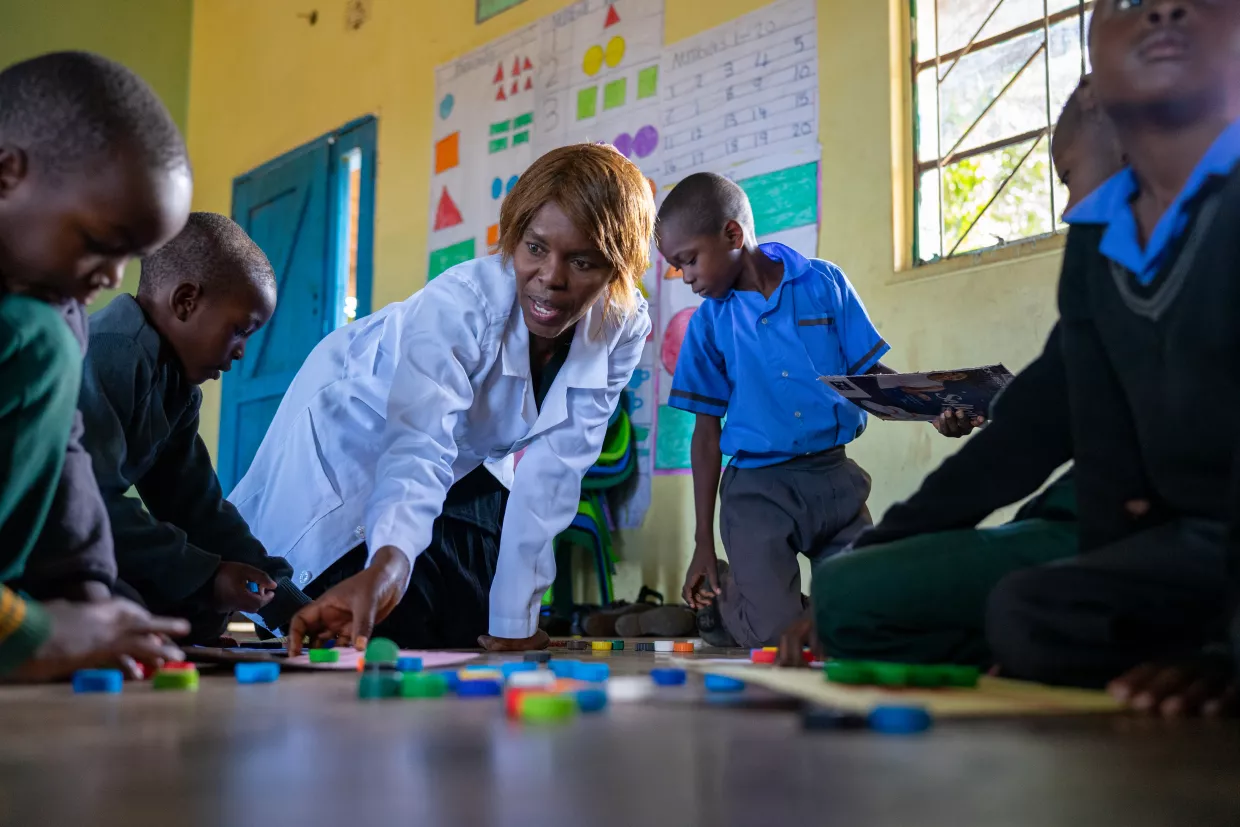 A teacher in a white coat enthusiastically engaging with children in a classroom, who work on crafts.