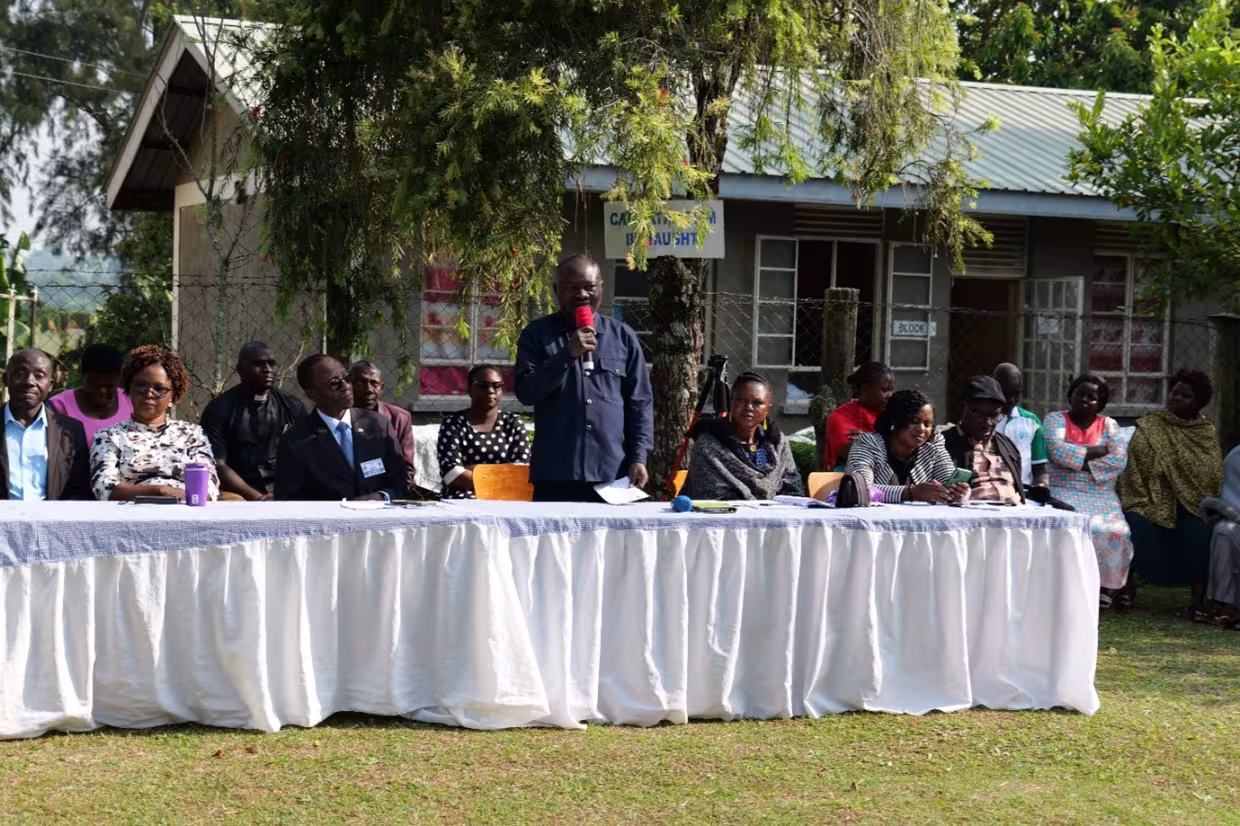 "A speaker addresses an audience outdoors during a school event, with several people seated at a long table