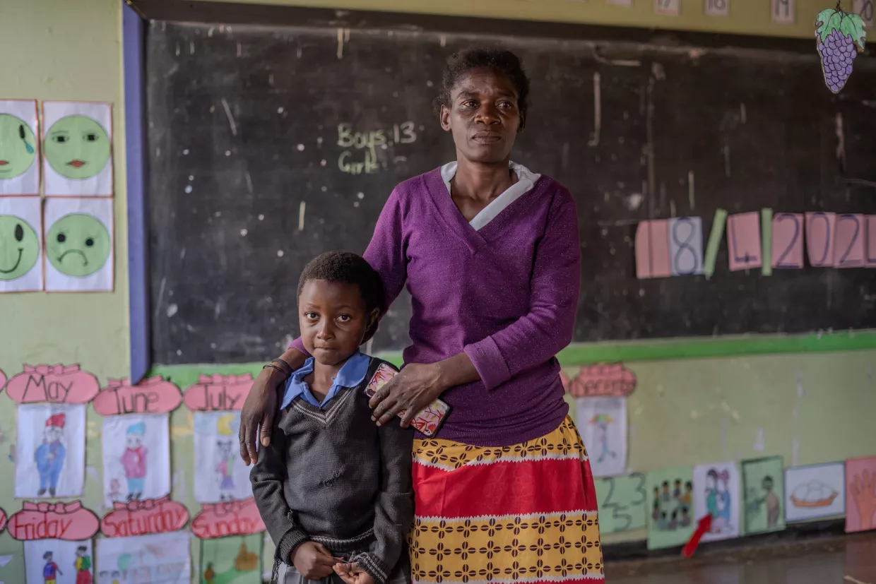 Jane stands beside her granddaughter Blessing in a colourful classroom, her arm resting gently on the her shoulder.
