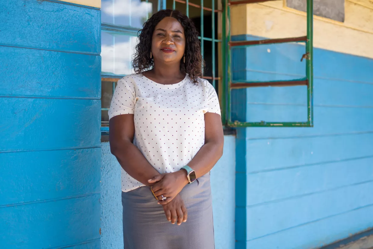 Teacher Emelda Musonda standing outside her classroom. Her left hand is holding her right arm. The walls are painted a bright blue.