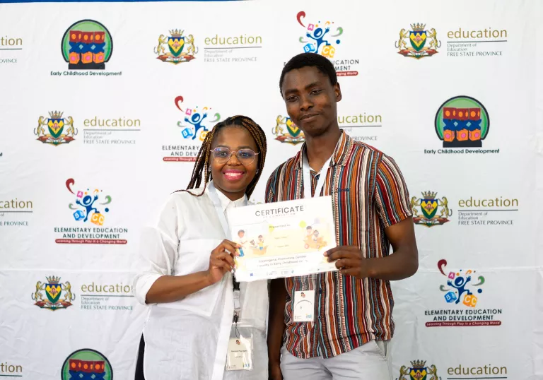 Two participants attending a celebration ceremony of teachers completing learning in gender-transformative pedagogy in South Africa hold up their certificate of completion.