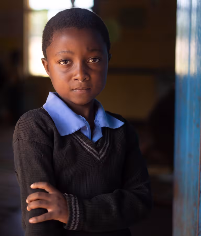 Blessings, a 6-year-old from Zambia is standing outside her classroom. She put one of her arms over the other looking directly into the camera lens.