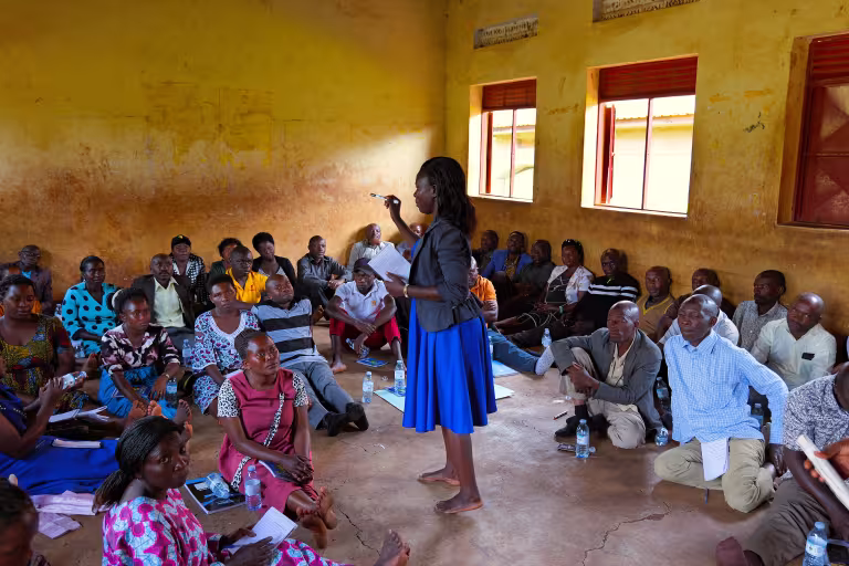 A lady standing and addressing a large seated group inside a classroom with yellowish walls, while participants sit on the floor listening and taking notes.