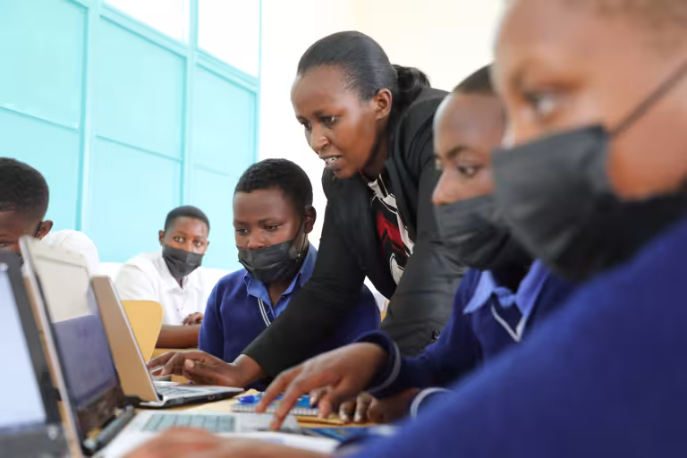 A teacher guides focused students wearing masks as they work on laptops in a bright classroom. The atmosphere is attentive and educational.