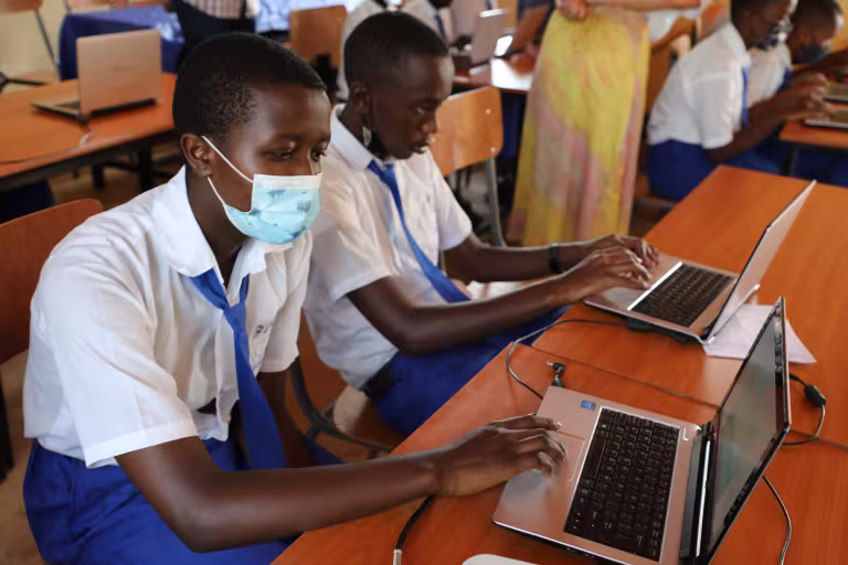 Two students in school uniforms work on laptops in a classroom. One wears a face mask. The scene conveys focus and engagement in a learning environment.