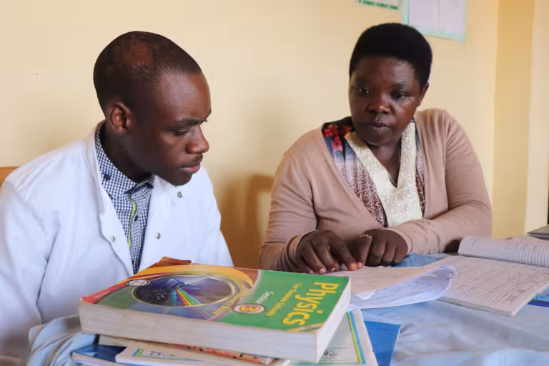 Female educator guiding a male trainee teacher during a study session, reviewing lesson plans and a physics textbook in a classroom setting, highlighting mentorship and teacher training.