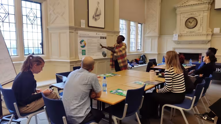 A woman presents a research poster to a small group seated around tables in a bright meeting room with tall decorative windows, blue chairs, and an ornate fireplace and clock.