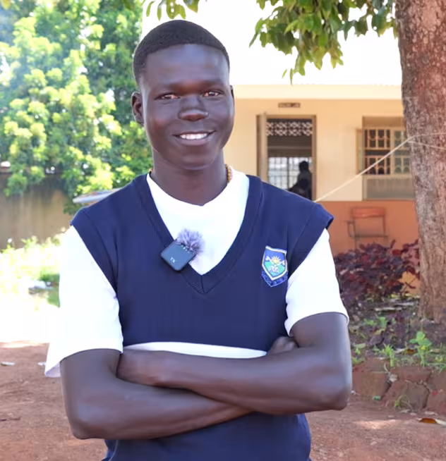 Smiling Black student in a school uniform stands outdoors with arms crossed, wearing a navy sweater vest and microphone in a sunny courtyard.