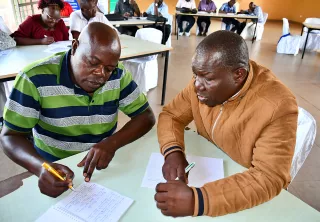 Two men engage in discussion while writing notes in a large room.