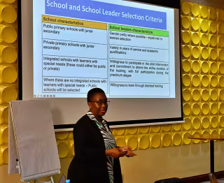 A person presents a slide on "School and School Leader Selection Criteria" in a conference room. 
