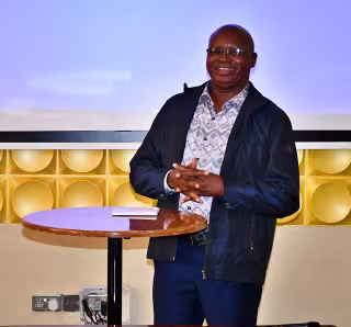 Smiling man in glasses, dark jacket, and patterned shirt, stands by a round wooden table with a notepad, in front of a bright projector screen.