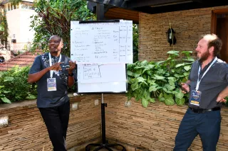 Two men stand outside by a whiteboard with diagrams, engaged in discussion.
