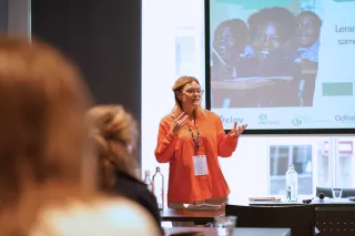 A woman in an orange shirt speaks confidently to an audience in a conference room. A projection screen behind her shows images of children learning.