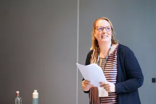 Smiling woman in glasses holds papers, standing against a plain background. She wears a striped shirt and cardigan, conveying a confident and positive tone.